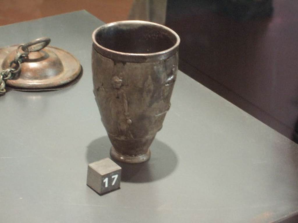 II.7.9 Pompeii. Palaestra. Found May 15 1936. Silver cup with scenes of the cult of Isis.
One of two (6044 and 6045) found near the body of a man in the Palaestra, possibly an initiate of the Isis cult.
The goddess is depicted here with her head facing backwards. 
Isis holds a sistrum in one arm, which is raised and bent, while with the other arm, held by her side, she holds a situla. 
Now in Naples Archaeological Museum. Inventory number 6044.

The second silver cup (6045), not pictured here, shows a bald priest is carrying a container (urnula), holding sacred water from the Nile, to the goddess Isis.
Isis carries in her bent arm a statuette of the Falcon god Seker or Sokaris, depicted standing on a small base. 
In front of her, on an altar, there is a vase upon which there is the god Horus. 
An arch supported by naked Caryatids, through which it is possible to see a series of steps with palms representing the sacred area, completes the scene. 
These two cups represent an example of sacred, rather than household, furnishings.
Now in Naples Archaeological Museum. Inventory number 6045.

