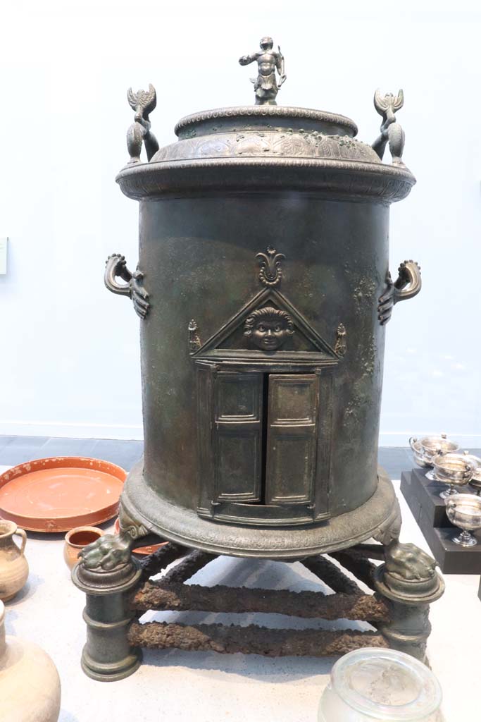 VIII.1.4 Pompeii. February 2021. 
According to Conticello (in Rediscovering Pompeii, p.173, no.59) –
This is a bronze cylindrical food warmer found under the stairs in the servant’s quarters of I.8.17.
Photo courtesy of Fabien Bièvre-Perrin (CC BY-NC-SA).
The Romans created a range of richly decorated devices, usually in bronze, for storing hot water during triclinium banquets. 
The hollow cylinder of the latter rests on an iron tripod. 
The feet are in the form of lion's paws, the handles end in small hands. 
A small temple with a Gorgon in the tympanum can be seen in the opening. 
Two dolphins and a Triton decorate the lid.
See Le Musée Maillol : Pompéi - Un art de vivre, l’exposition du 21 septembre 2011 au 12 février 2012. Communiqué de presse.
Parco Archeologico Pompei, inventory number 6798.
