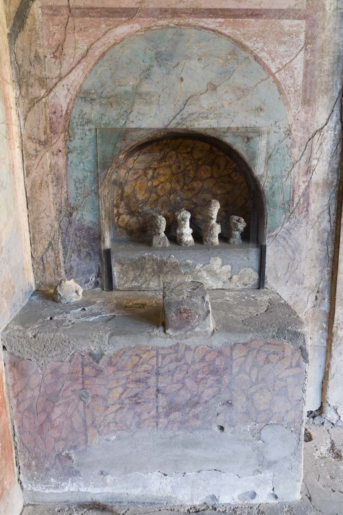 I.10.4 Pompeii. April 2022.
Alcove 25, looking towards altar to household gods against west wall. Photo courtesy of Johannes Eber.
According to Boyce –
In the finely decorated exedra opening off the south-west corner of the peristyle is an unusual shrine.
A large rectangular, masonry podium (1.33 by 0.66, h.089) is built into the south-west corner.
In the west wall above it is a shallow arched niche (h.0.62, w.0.86, h. above the podium 0.24), set into the centre of a previously existing panel of the wall decoration. A small, rectangular, altar-like base of tufa (0.21 by 0.17, h.0.25) was built directly before the niche on the top of the podium.
The large podium is painted to represent a veneering with vertical slabs of yellow marble veined with red, placed above a dark dado.
The back wall of the niche is painted in imitation of the same marble.
The small altar-like structure is coated with red stucco.
Within the niche there stood in antiquity five figures of perishable material which left their impressions in the volcanic ash; of these impressions the excavators were able to take plaster casts. ………….
See Boyce G. K., 1937. Corpus of the Lararia of Pompeii. Rome: MAAR 14. (p.28.no. 49B).
See Giacobello, F., 2008. Larari Pompeiani: Iconografia e culto dei Lari in ambito domestico. Milano: LED Edizioni, (p.256, no.V12)