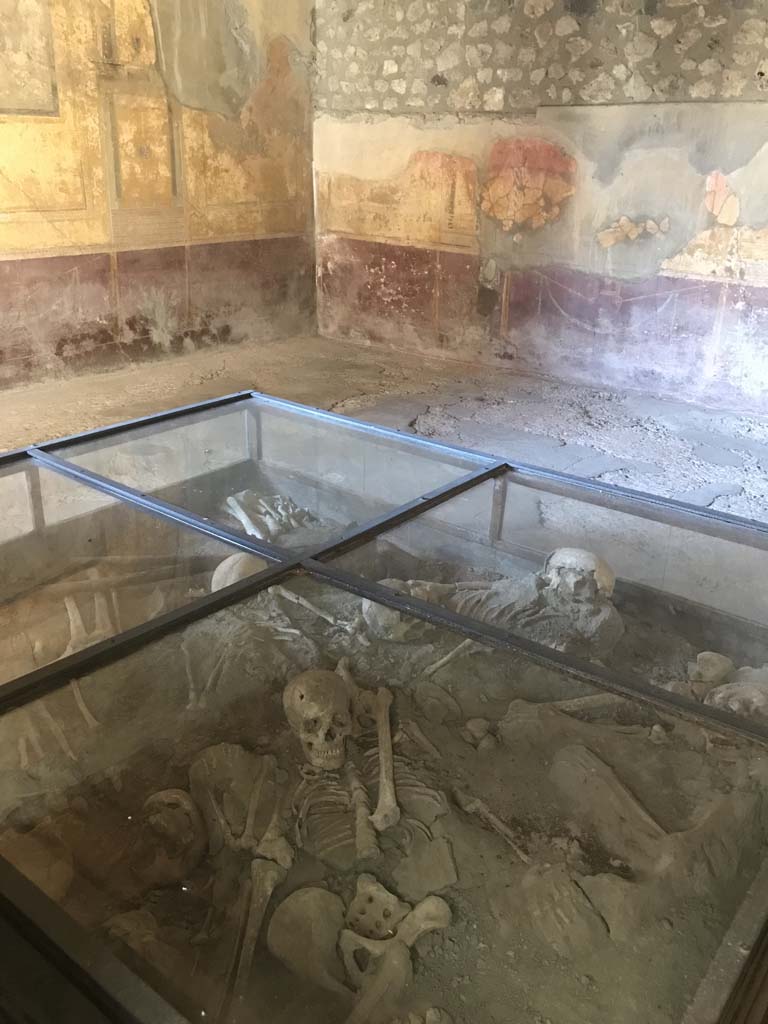 I.10.4 Pompeii. April 2019.
Room 19, looking towards north-east corner. Photo courtesy of Rick Bauer.
On one wall is a painting of a satyr playing a flute or pipes to a maenad.
In this room is also a wall painting of a cherub scared by a tragic mask held by a nymph.
Three skeletons (which were removed) were found here with a mattock and pickaxe which may have been used to dig the holes in the walls of the room to try to escape.
The display case contains skeletons of 10 others, who were found a few days later in the adjacent corridor P1 at about 2.5 metres above the ground.
They appeared to have fallen from an upstairs room.