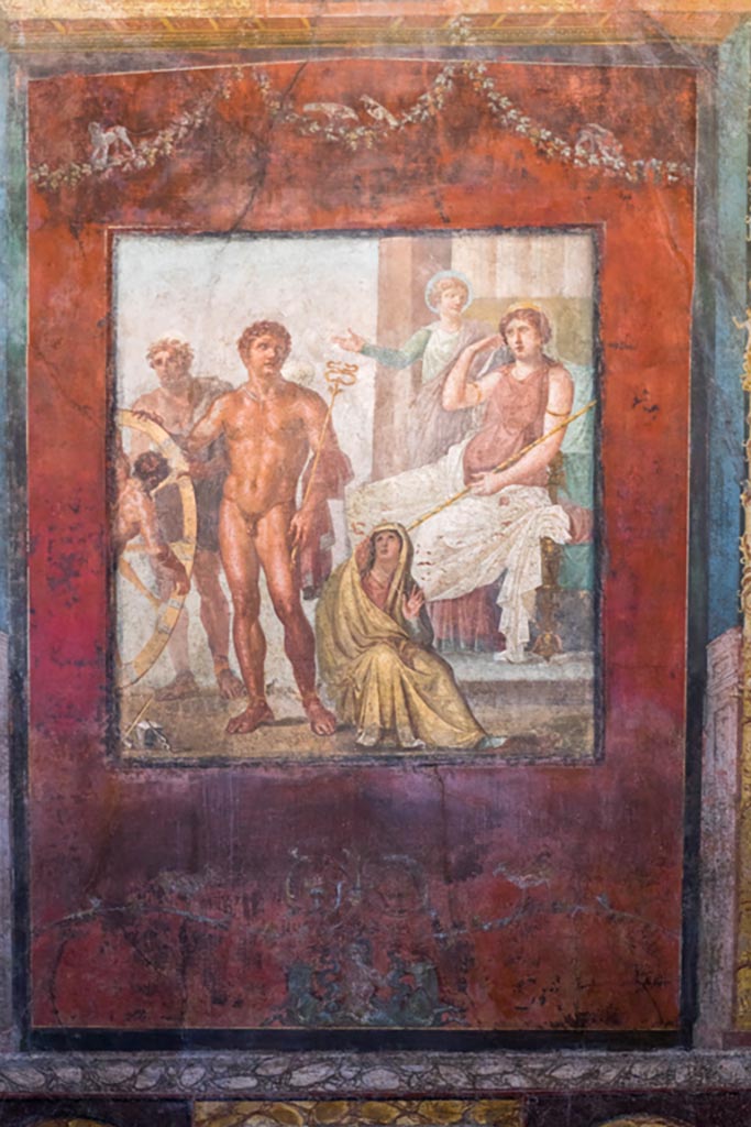 VI.15.1 Pompeii. March 2023. 
Central wall painting from east wall of exedra - the torture of Ixion on the wheel. 
Photo courtesy of Johannes Eber.
Ixion has already been attached to the wheel by Hephaestus.
Hermes/Mercury stands to the right with his caduceus, helmet and sandals.
The figure seated on the floor is perhaps the cloud nymph Nephele or a personification of the underworld.
Hera/Juno is seated on her throne with Iris standing to her left, gesticulating.
See Carratelli, G. P., 1990-2003. Pompei: Pitture e Mosaici: Vol. V. Roma: Istituto della enciclopedia italiana, p.538. 
