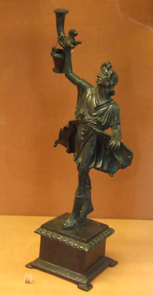 Bronze statuette of a Lar, found in Lararium of VI.16.7.
This was one of two Lares that were found on the lower step of the lararium.
The Lar holds a rhyton which has at its lower end the front half of a panther.
The bucket would normally be in the Lar’s left hand.
Now in Naples Archaeological Museum. Inventory number 133328.
See Notizie degli Scavi di Antichità, 1907, p. 567, fig. 17.