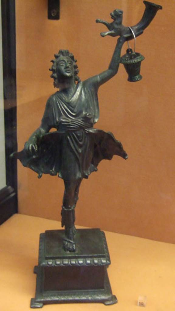 Bronze statuette of a Lar, found in Lararium of VI.16.7.
This was one of two Lares that were found on the lower step of the lararium.
The Lar holds a rhyton which has at its lower end the front half of a panther.
The bucket would normally be in the Lar’s right hand.
Now in Naples Archaeological Museum. Inventory number 133327.
See Notizie degli Scavi di Antichità, 1907, p. 567, fig. 17.