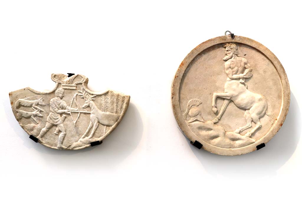 VI.16.7 Pompeii. February 2021. On right: Decoration (oscilla) hung between columns in the porticoes, showing centaur.
On the left from I.4.25 is a pelta showing scenes of Hercules’ life.
Photographed in Antiquarium. Photo courtesy of Fabien Bièvre-Perrin (CC BY-NC-SA).