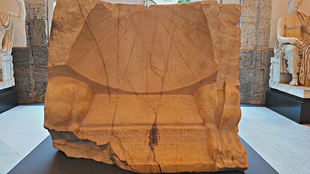 VII.1.8 Pompeii. November 2018.
Marble sundial with Oscan inscription, found 23rd September 1854. Photo courtesy of Giuseppe Ciaramella.
An inscription in Oscan reveals that the quaestor Maras Atinius commissioned the construction of the sundial:
“Maras Artinius (son of) Maras, quaestor, with the money from the fines, by resolution of the assembly, had (this) made”.
Solar clocks were one of the many contributions of the Greeks to the Roman world, and this refined Pompeian specimen bears witness to the Hellenistic taste to which the Terme Stabiane (a modern name referring to their position along the Via Stabiana) conformed. The Terme Stabiane replaced an earlier, much less sophisticated facility in the 2nd century BC.
Discovered at Pompeii on 23 September 1854 in the Stabian Baths, it was used to regulate the turns for entering the complex.
The dial is a portion of a right circular cone, bounded on the upper side by a hyperbola.
The clock has three circular day curves that are parallel to each other.
The day curves are the lines cast by the shadow of the gnomon over the course of a whole day. The arcs of the solstices and equinoxes are each divided into 12 equal parts to plot the eleven hour lines, obtained by joining the corresponding partitions of the three day curves.
The hours marked are “unequal hours”.
Now in Naples Archaeological Museum. Inventory number 2541.