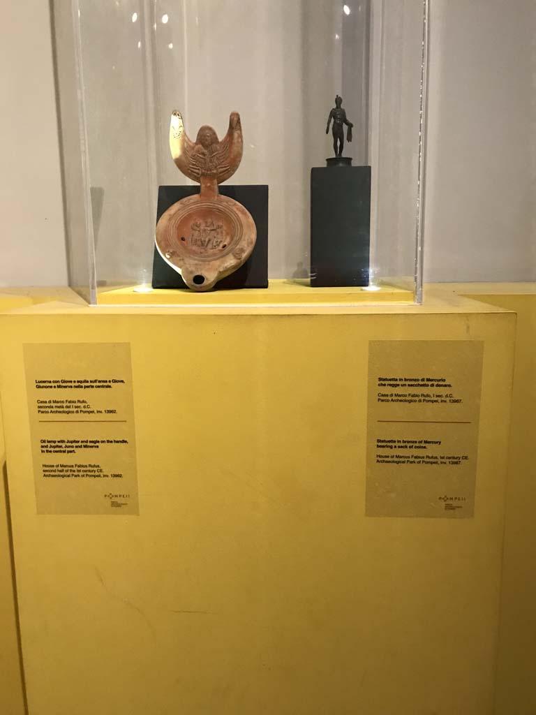 VII.16.17-22, Pompeii. House of Fabius Rufus. April 2019. Lamp and statuette. In exhibition in Antiquarium.
Oil lamp with Jupiter and eagle on the handle, and Jupiter, Juno and Minerva in the central part. PAP inventory number 13962.
Bronze statuette of Mercury carrying a sack of coins. PAP inventory number 13987.
Photo courtesy of Rick Bauer.