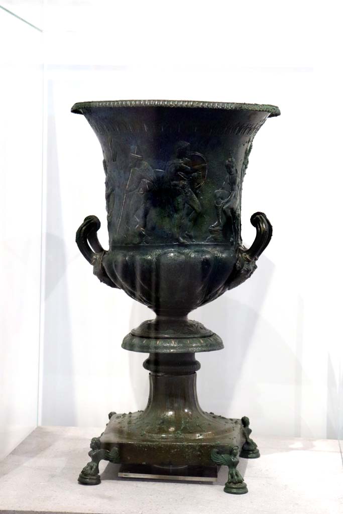 VIII.1.4 Pompeii. February 2021. 
Bronze krater with scenes in relief, found in the triclinium at IX.13.1-3, on display in Antiquarium.
Photo courtesy of Fabien Bi�vre-Perrin (CC BY-NC-SA).
According to the description card in the Antiquarium -
The bronze krater was decorated with eight armed male figures, six standing and two sitting. 
One of the seated figures is tying his shoelaces.
A young man stands in front of him holding a shield, with a bearded figure holding a staff behind.
Young and naked, the second seated figure caresses an animal with one hand and holds a sword by the hilt in the other.
(See IX.13.1-3 for more photos).
