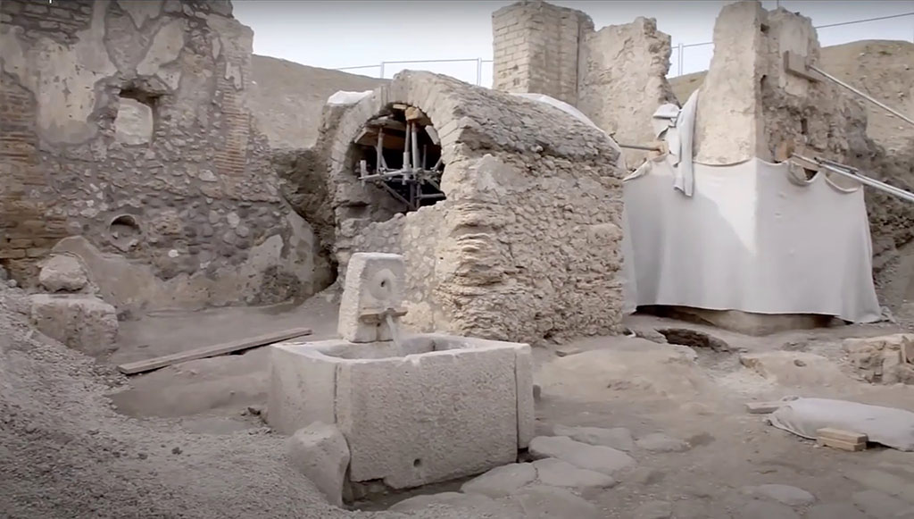 Pompeii Fountain at V.8. Fountain at crossroads of Vicolo delle Nozze d’Argento and Vicolo dei Balconi.
The newly discovered fountain is in a small square open area with a well/cistern with arched roof behind and a water tower at rear. 
The bar with the gladiator fresco is on the back of the wall to the right of the water tower and behind the well/cistern.

Pompei V.8. Fontana al quadrivio di Vicolo delle Nozze d'Argento e Vicolo dei Balconi.
La fontana appena scoperta si trova in una piccola area quadrata aperta con un pozzo / cisterna con tetto ad arco dietro e una torre d'acqua sul retro. 
Il bar con l'affresco gladiatore si trova sul retro della parete a destra della torre dell'acqua e dietro il pozzo /cisterna.

Photograph © Parco Archeologico di Pompei.
