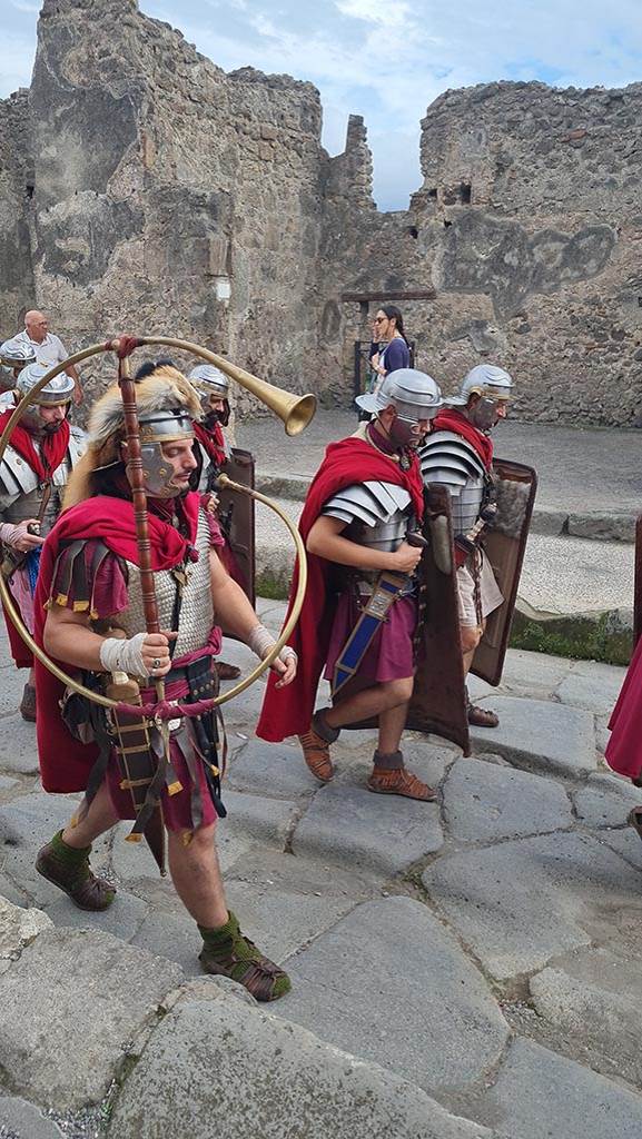 I.4.17 Pompeii. 28th September 2024 during “Ludi Pompeiani” event.  
Looking south across Via dell’Abbondanza. 
Roman soldiers marching including the “Cornicen” holding a C shaped brass instrument (cornu) used to signal orders to Legions.
Photo courtesy of Giuseppe Ciaramella.

