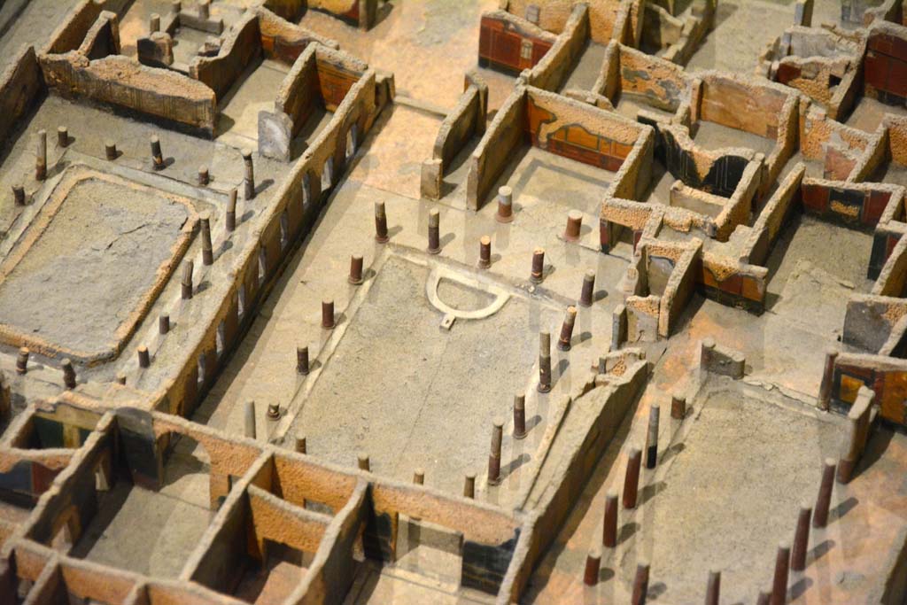 I.4.25 Pompeii. May 2019. Detail from model in Naples Archaeological Museum.
Looking south-west across Middle Peristyle 17, in centre. 
Foto Tobias Busen, ERC Grant 681269 DÉCOR

According to Jashemski –
“The centre garden, (the Middle Peristyle), was surrounded by a portico supported by seventeen columns made of brick and Sarno limestone and faced with stucco. The upper parts were fluted, the bottoms left plain and painted red. There was a gutter around the edge of the garden. 
At the west end of the garden was a semi-circular basin around which were arranged six spectacular bronze animals found in 1861.  …………………………
In February 1926, when restoring the garden in this peristyle, Maiuri discovered a large rectangular pool (3.50m wide x 10.80m long x 1.40m deep). On the west side two high steps (0.45m and 0.40m) led down to the pool. The rough foundations of the much smaller semi-circular pool had been built on these steps and on the bottom of the large pool. The large pool had then been filled with old building material and soil. This was probably at the time that the house with the entrance on the Via dell’Abbondanza was joined to this house, probably after the earthquake. The cistern opening in the south-west corner of the garden had a lava puteal; the one in the east portico, no puteal.”
See Jashemski, W. F., 1993. The Gardens of Pompeii, Volume II: Appendices. New York: Caratzas. (p.29-30).
