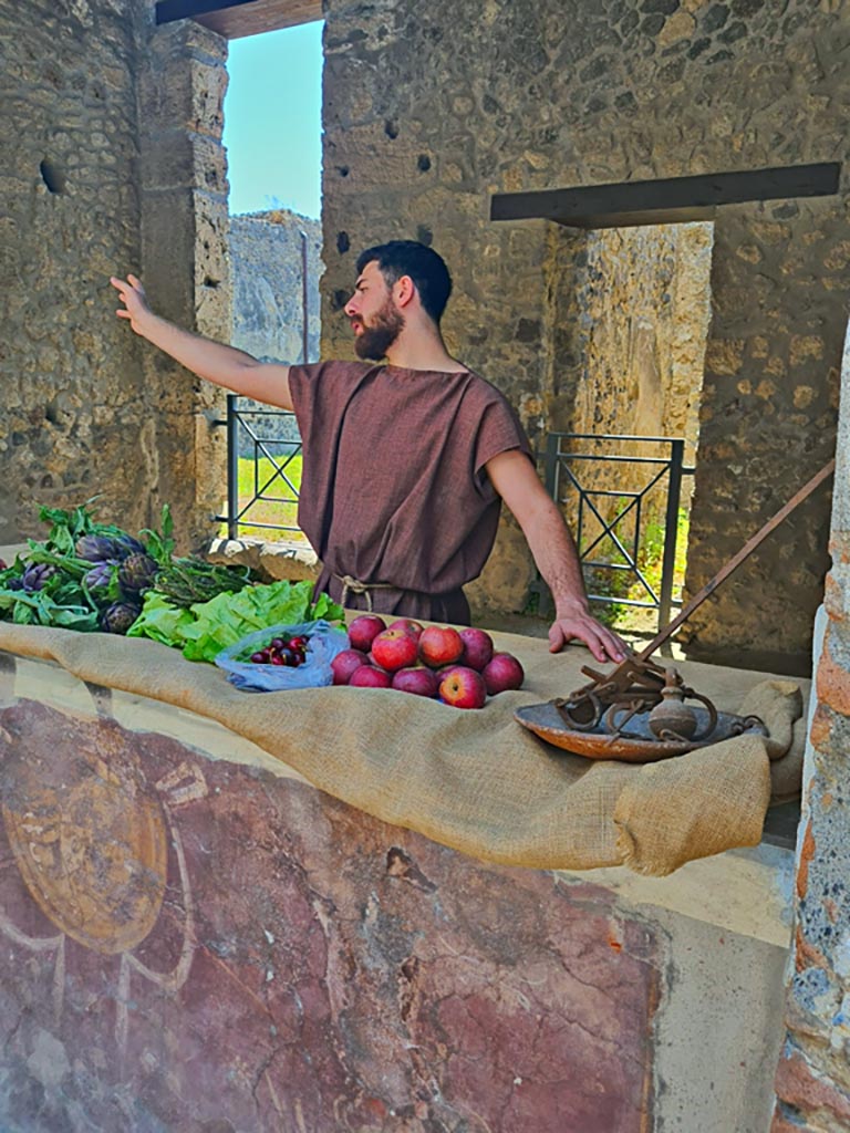 I.8.1 Pompeii. 8th June 2024. 
Looking south-east across counter, with actor playing part of ancient pompeian advertising his wares to the customers. 
Photo courtesy of Giuseppe Ciaramella.
Historical reconstruction entitled L’altra Pompei prende vita (The other Pompeii comes to life).
