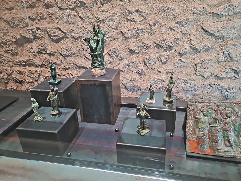 II.7.9 Pompeii. Palaestra. March 2024.
Items in display case in exhibition entitled – “L’altra Pompei, vite comuni all’ombra del Vesuvio”. Photo courtesy of Giuseppe Ciaramella.
No.1, 2 and 3, on right – from the Isiac Lararium at Civita Giuliana, Imperial Villa.
No. 4, on left, Statuettes of Isis/Fortuna, from Villa della Pisanella, Boscoreale.
No. 5, centre rear – Hand of Sabazios, from Magic Rites Complex, II.1.12 Pompeii.
No. 6, rear left – Statuette of Attis, from II.1.3 Pompeii.