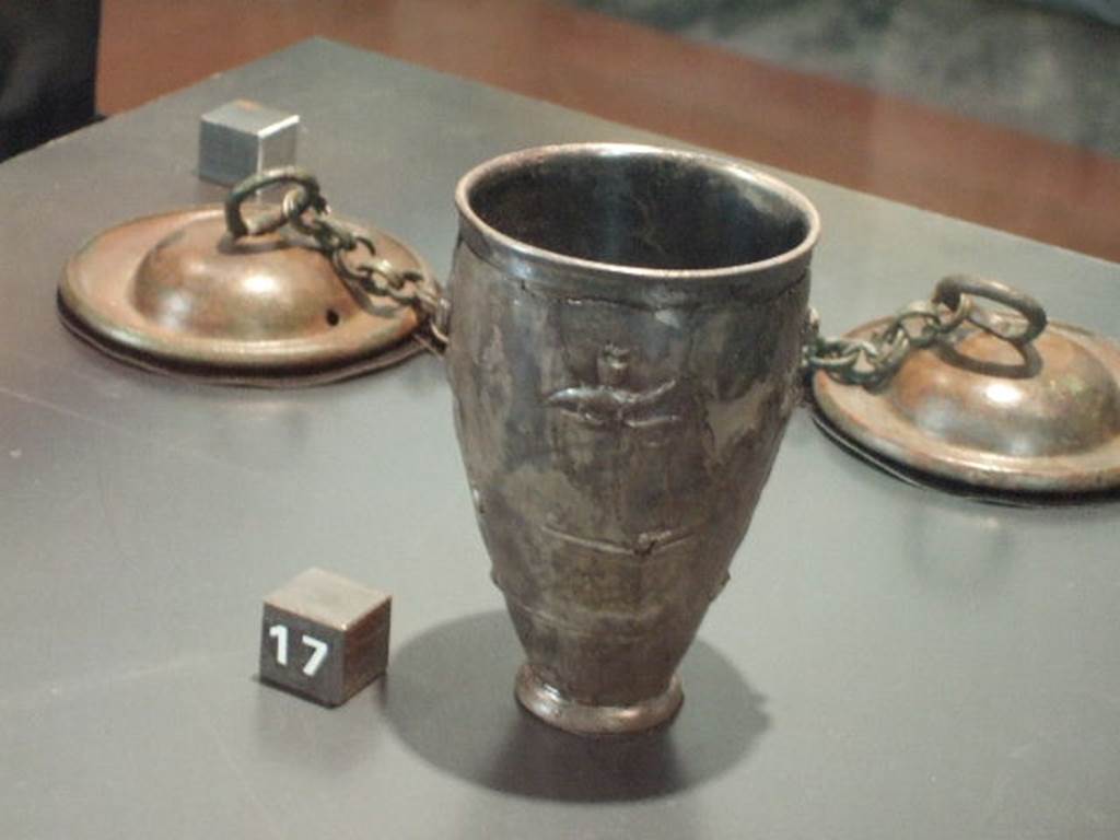 II.7.9 Pompeii. Palaestra. Silver cup with scenes of the cult of Isis. One of two found near the body of a man in the Palestra, possibly an initiate of the Isis cult. According to the Naples Archaeological Museum web site, the scene depicts the cult of Isis. A bald priest is carrying a container for holding sacred water from the Nile to the goddess Isis. Isis carries in her bent arm a statuette of the Falcon god Sokaris, depicted standing on a small base. In front of her, on an altar, there is a vase upon which there is the god Horus. An arch supported by naked Caryatids, through which it is possible to see a series of steps with palms representing the sacred area, completes the scene. These two cups represent an example of sacred, rather than household, furnishings. Now in Naples Archaeological Museum. Inventory number 6045.