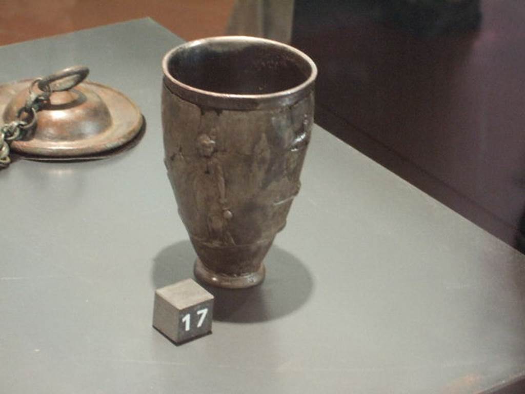 II.7.9 Pompeii. Palaestra. Found May 15 1936. Silver cup with scenes of the cult of Isis.
One of two (6044 and 6045) found near the body of a man in the Palaestra, possibly an initiate of the Isis cult.
The goddess is depicted here with her head facing backwards.
Isis holds a sistrum in one arm, which is raised and bent, while with the other arm, held by her side, she holds a situla.
Now in Naples Archaeological Museum. Inventory number 6044.
The second silver cup (6045), not pictured here, shows a bald priest is carrying a container (urnula), holding sacred water from the Nile, to the goddess Isis.
Isis carries in her bent arm a statuette of the Falcon god Seker or Sokaris, depicted standing on a small base.
In front of her, on an altar, there is a vase upon which there is the god Horus.
An arch supported by naked Caryatids, through which it is possible to see a series of steps with palms representing the sacred area, completes the scene.
These two cups represent an example of sacred, rather than household, furnishings.
Now in Naples Archaeological Museum. Inventory number 6045.