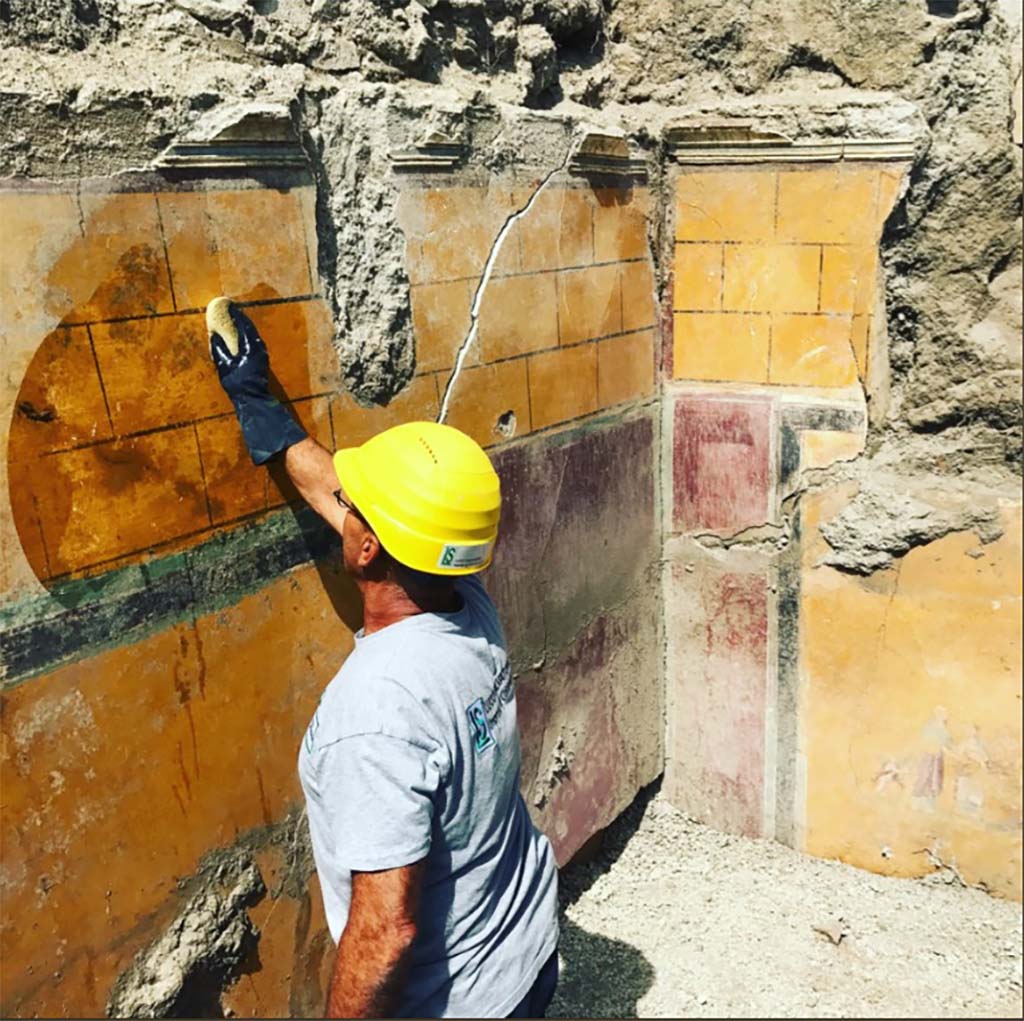 V.2.21 Pompeii. May 2018. Room A9 (as shown on PAP plan), north-west corner. On the north wall is a painting of figures with a cupid.
It is most likely a cubiculum (bedroom), due to the presence on one side of the recess intended to house the headboard of the bed.
Si trattava molto probabilmente di un cubiculum (stanza da letto), per la presenza su un lato dell’incasso destinato a ospitare la testata del letto. 
See http://pompeiisites.org/wp-content/uploads/Cartella-Stampa-Regio-V-8-gugno.docx 
Photograph Massimo Osanna © Parco Archeologico di Pompei.
