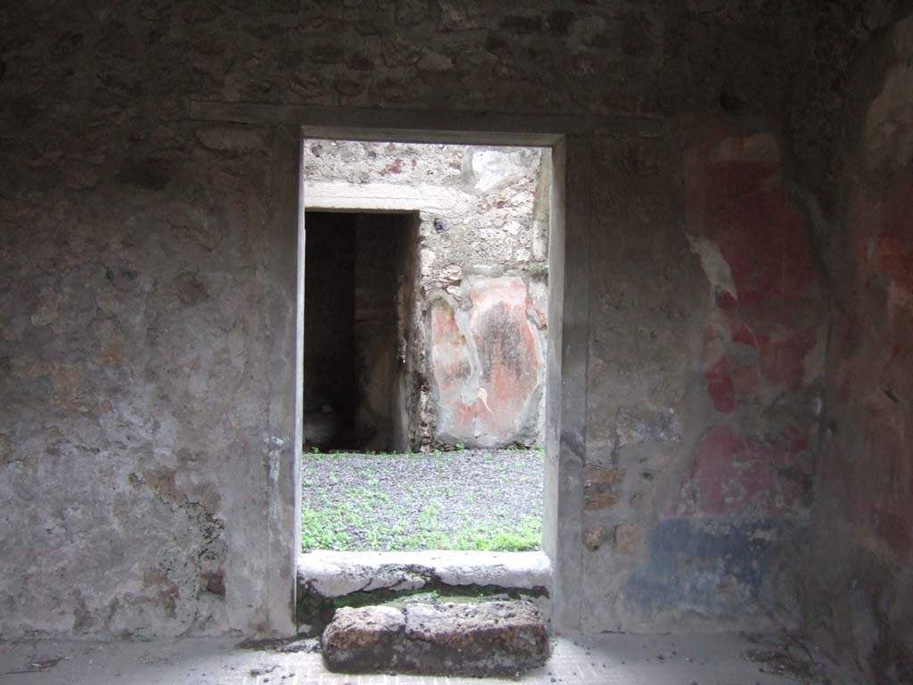 V.2.h Pompeii. December 2005. South wall of room ‘f’ the tablinum, with doorway to rear.     
Opposite the entrance fauces was the tablinum, situated between the corridor and the cubiculum.
The tablinum had flooring of signinum and walls with a red background, with black dado and white frieze.
The decoration had nearly all fallen from the south wall.
In the south wall was a block of limestone, which served instead of a step to the doorway which opened out into the small garden ‘h’.
In the central panels of the east and west walls were the circular paintings with heads of youths with crowns of foliage.
These are now found in the Naples Archaeological Museum, (see NdS, 1892, p.28)
Another two medallions with heads were painted on the south wall near to the above-mentioned doorway, as could be seen by the remains of a medallion on the pilaster to the right, in which part of the head was visible.
In the side panels were medallions with landscapes.
Also, here and there on the walls of the tablinum could be seen traces of fire.
See Notizie degli Scavi di Antichità, 1896, (p.422) which refers to tablinum ‘b’, corridor ‘c’, cubiculum ‘d’ and the garden ‘f’.
