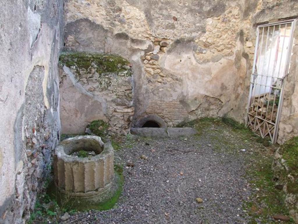 V.2.i Pompeii.  December 2007.  Room 11.  Kitchen.  Wall at south end near the caldarium showing a hole in the floor, used to heat the bath rooms.  On the left in the south east corner are the remains of a masonry foundation, possibly to support the lead vessel in which water was kept for the bath.  The stone puteal in the form of a Doric column does not cover a cistern mouth.  See Allison, P. M. Pompeian Households: An online companion.  http://www.stoa.org/projects/ph/rooms?houseid=16#293 (room s).  On the right on the west wall is the door (now gated) to the latrine.