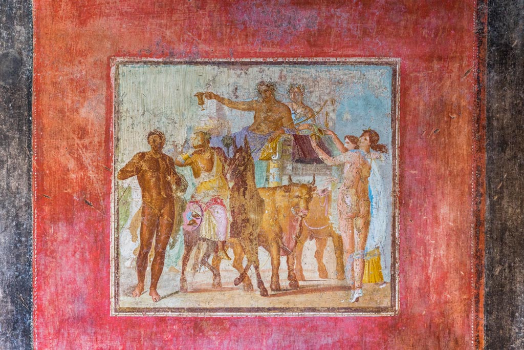 V.4.a Pompeii. January 2023. 
Room ‘h’, central red panel with wall painting of Bacchus and Ariadne riding on a chariot drawn by two oxen
Photo courtesy of Johannes Eber.
Kuivalainen comments –
“This is the high point of the thiasus; all the participants are depicted in their colourful finery, and music is played in the procession. The artist shows a considerable level of skill in presenting his figures from many different viewpoints and with foreshortenings in a tight composition. Ariadne is depicted taking care of the intoxicated Bacchus. Silenus is riding a donkey, as in the wall painting in the Casa delle Amazzoni, B16.”
See Kuivalainen, I., 2021. The Portrayal of Pompeian Bacchus. Commentationes Humanarum Litterarum 140. Helsinki: Finnish Society of Sciences and Letters, (p.135-36, D14).

