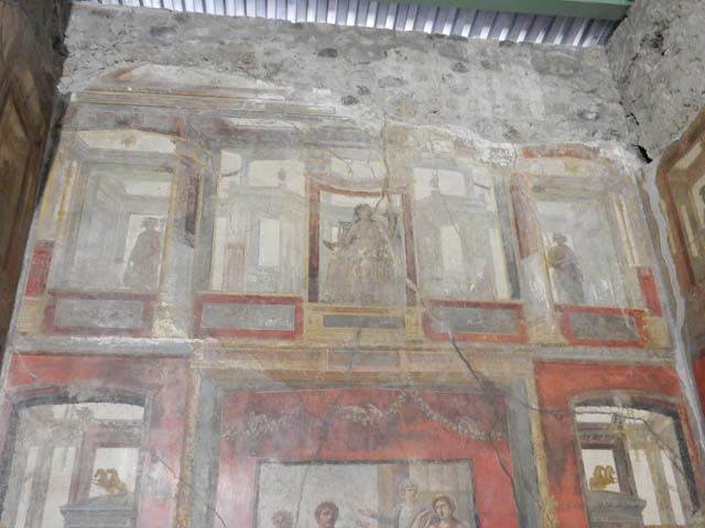 VI.15.1 Pompeii. May 2017. Upper south wall of the exedra. Photo courtesy of Buzz Ferebee.
In the upper centre of the south wall, damaged, a figure with left shoulder destroyed, and fractures throughout the painting. 
Kuivalainen describes – 
“Inside an aedicula on a white background, a youth sits on a throne with his legs on a footstool: he leans with his left hand on a long thyrsus and wears high boots and a red cloak on his hips: the yellowish legs of the throne are turned. 
Kuivalainen comments –
“The whole south wall is thematically Bacchic, and this young half-naked Bacchus is depicted among other gods in the architecture of the upper zone.”
See Kuivalainen, I., 2021. The Portrayal of Pompeian Bacchus. Commentationes Humanarum Litterarum 140. Helsinki: Finnish Society of Sciences and Letters, (p.100-1, B11).
