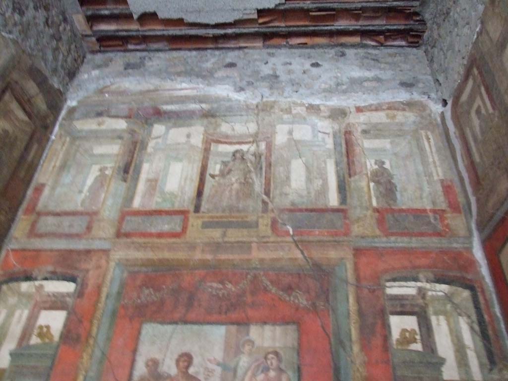 VI.15.1 Pompeii. January 2017. Looking towards upper south wall of the exedra.
Foto Annette Haug, ERC Grant 681269 DÉCOR.
In the upper centre of the south wall, damaged, a figure with left shoulder destroyed, and fractures throughout the painting. 
Kuivalainen describes – 
“Inside an aedicula on a white background, a youth sits on a throne with his legs on a footstool: he leans with his left hand on a long thyrsus and wears high boots and a red cloak on his hips: the yellowish legs of the throne are turned. 
Kuivalainen comments –
“The whole south wall is thematically Bacchic, and this young half-naked Bacchus is depicted among other gods in the architecture of the upper zone.”
See Kuivalainen, I., 2021. The Portrayal of Pompeian Bacchus. Commentationes Humanarum Litterarum 140. Helsinki: Finnish Society of Sciences and Letters, (p.100-1, B11).

