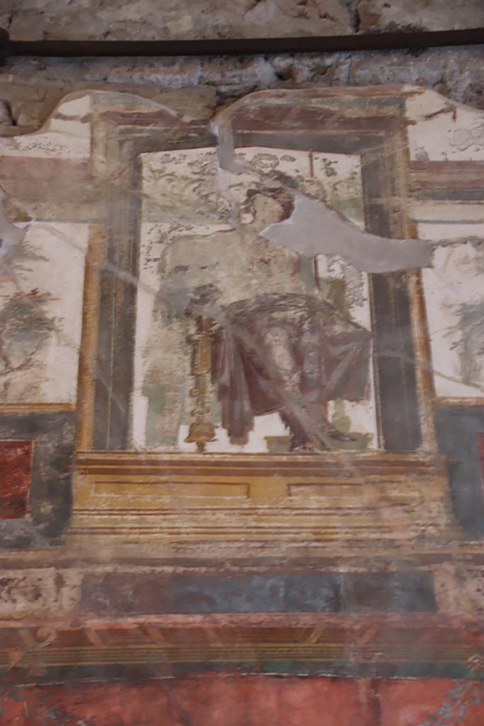 VI.15.1 Pompeii. October 2023. 
Upper south wall, painted seated central figure from above central panel. Photo courtesy of Klaus Heese.
In the upper centre of the south wall, damaged, a figure with left shoulder destroyed, and fractures throughout the painting. 
Kuivalainen describes – 
“Inside an aedicula on a white background, a youth sits on a throne with his legs on a footstool: he leans with his left hand on a long thyrsus and wears high boots and a red cloak on his hips: the yellowish legs of the throne are turned. 
Kuivalainen comments –
“The whole south wall is thematically Bacchic, and this young half-naked Bacchus is depicted among other gods in the architecture of the upper zone.”
See Kuivalainen, I., 2021. The Portrayal of Pompeian Bacchus. Commentationes Humanarum Litterarum 140. Helsinki: Finnish Society of Sciences and Letters, (p.100-1, B11).

