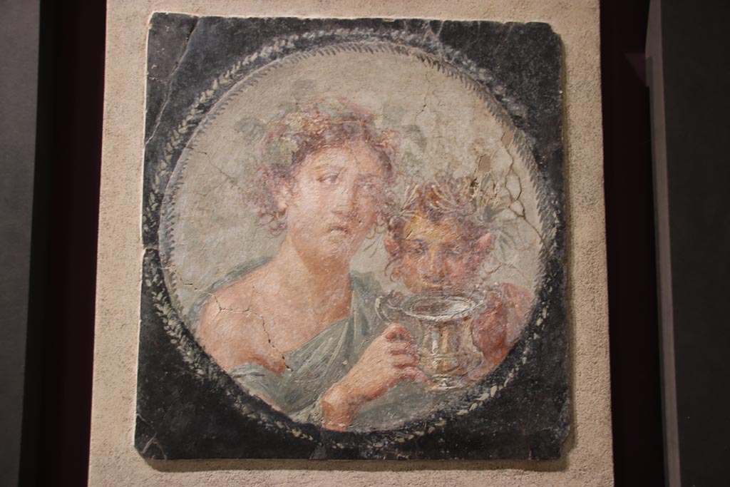 VI.17.42 Pompeii. October 2022. Medallion depicting Maenad and a young Satyr holding a silver drinking cup for wine (a kantharos).
Found at the west end of the north wall of the large triclinium with a vaulted ceiling. On display in exhibition in Palaestra.
Photo courtesy of Klaus Heese.