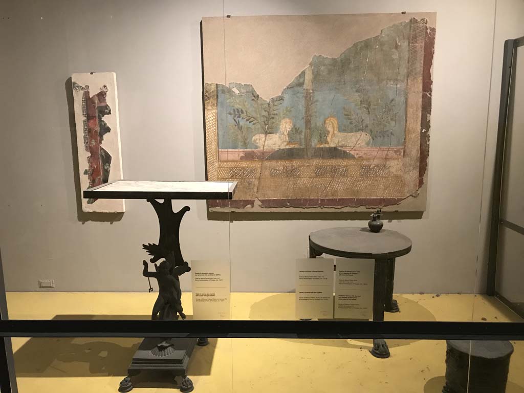 VII.16.17-22 Pompeii. April 2019. On display in Antiquarium, from House of Fabius Rufus.
Bronze table with cupid riding a dolphin and marble top. PAP inventory number 13371.
Bronze table with lion's feet. PAP inventory number 13108.
On table is a bronze pitcher with face of a follower of Dionysus. PAP inventory number 14072.
Fragment of fresco on the wall at the rear, from VI.17.42, Pompeii.
Fragment of the wall of the summer triclinium 31 decorated with a pair of facing Sphynxes within a lush garden.
Parco Archeologico di Pompei, inventory number 87228.
Photo courtesy of Rick Bauer.