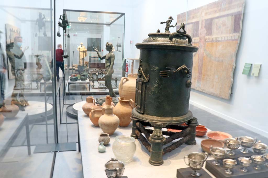 VIII.1.4 Pompeii. February 2021.
Looking across display cabinet containing silver from Moregine and bronze food warmer from I.8.17 (this end), and at the far end –
Bronze strainer and two pitchers from new excavations in Reg. V,
and Terracotta bottles, jugs and red ware plates and bowls from room 11, of the House of Orion, V.2.
Looking towards the bronze Ephebus lamp holder from VII.16.19-22, and across to the marble statue of Nike, from Oplontis, at the far end.
On the wall to the right is part of a painted wall from I.6.2.
Photo courtesy of Fabien Bièvre-Perrin (CC BY-NC-SA).