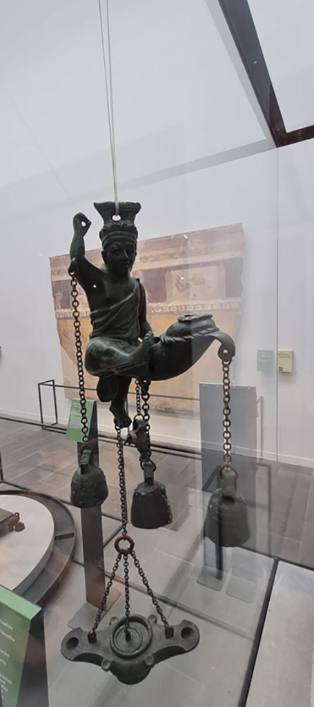 VIII.1.4 Pompeii. April 2022. Tintinnabulum.
Bronze lamp with statuette of a satyr with a phallus from which small bells hang, found in I.6.3.
Photo courtesy of Giuseppe Ciaramella.