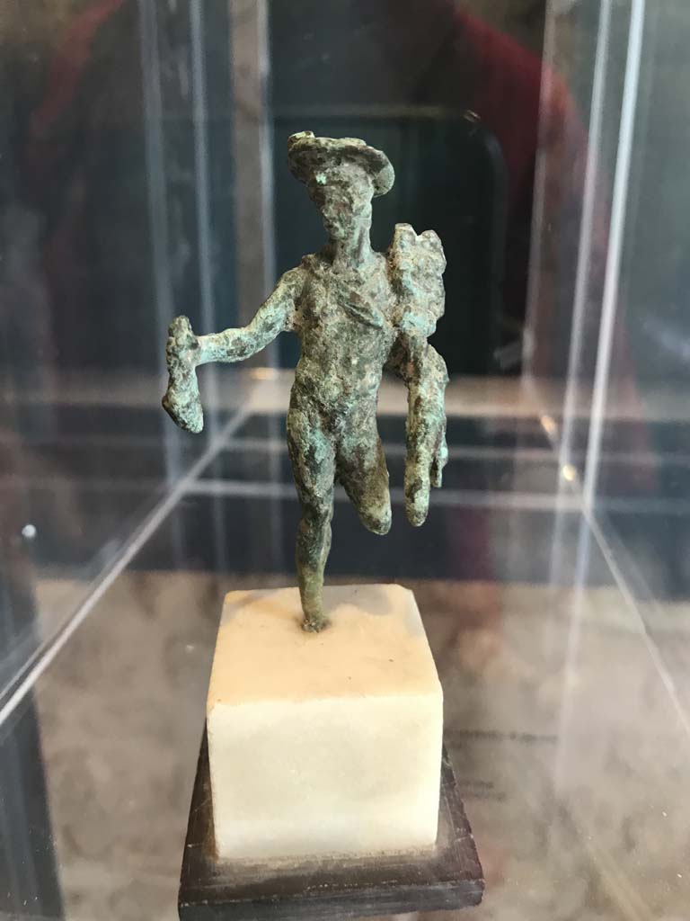 VIII.2.1 Pompeii. April 2019. Bronze statuette of Mercury carrying a money bag.
Found in the area to the south of the House of Championnet.
Pompeii Inventory number 6331. Photo courtesy of Rick Bauer.
According to Zanella -
This statuette was found in Room B, the triclinium on the lower floor of VIII.2.3-5 (or VIII.2.14-16) on 9th February 1937.
9/2/1937
In room B an imprint of a box had been removed at a distance of m. ,0,70 from the bed frame, this imprint had been found to be m.0.68 x 0.50 x 0.22 including a shelf in the front m.0.50 x 0.18 x 0.16.
In removing the last clod of ash with the partial imprint of the triclinium couch, the following was found: Bronze: statue representing Mercury standing missing a (leg) with the pouch and caduceus; it was fragmented and missing its left leg from the knee down and the right foot.
Alt m.0.068 (Pompeii inv. 6331)
(Nel ambiente B si era rimosso un’impronta di cassa distante m. ,0,70 dal telaio del letto, detta impronta era risultata di m.0.68 x 0.50 x 0.22 compreso un ripiano nella parte anteriore m.0.50 x 0.18 x 0.16.
Nel rimuovere l’ultima zolla di cenere con l’impronta parziale del letto tricliniare si era rinvenuto: bronzo: statuina rappresentante Mercurio stante mancante di un (*) col marsupio e caducco; era frammentata e mancante della gamba sinistra dal ginocchio in giù e del piede destro. Alt m.0.068 (Pompei inv. 6331))
See Zanella S., 2019. La caccia fu buona: Pour une histoire des fouilles à Pompéi de Titus à l’Europe. Naples : Centre Jean Bérard, p. 268.