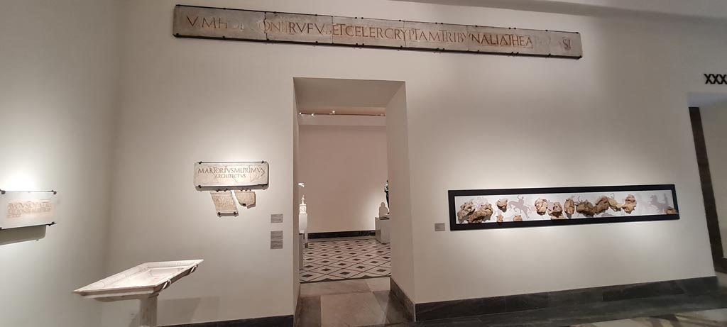 VIII.7.20 Pompeii. April 2023. 
Above doorway, (inv. 3821) one of the two identical white marble slabs with inscriptions each well over 6 metres long, from the area of the stage.
Left side of doorway, two marble slabs (inv. 3834 and 3823).
No. 3834 provides the name of the architect involved in the refurbishment of the theatre – M. Artorius Primus freedman of Marcus.
No. 3823 is a white marble slab in two parts, stating that M. Holconius Rufus and M. Holconius Celer arranged at their own expense for the enlargement of the seating and tribunal of the theatre and the construction of a vaulted corridor.
On display in “Campania Romana” gallery in Naples Archaeological Museum.  Photo courtesy of Giuseppe Ciaramella.
