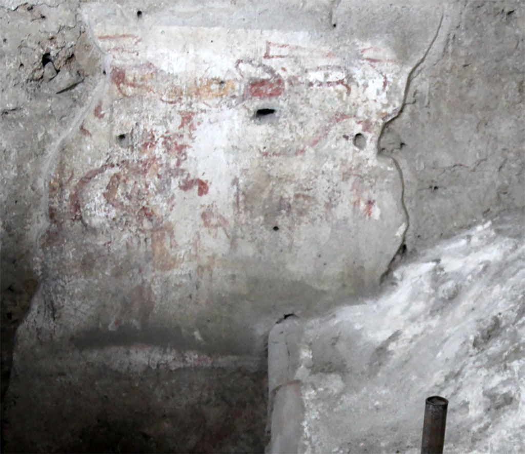 IX.12.B/IX.12.9 Pompeii. May 2023. Room “a”, south wall of kitchen on west side.
A male figure with a short tunic and cornucopia, interpretable as a lar.
Ambiente 13, parete sud della cucina sul lato ovest.
Una figura maschile con tunica corta e cornucopia, interpretabile come lare.
Photograph © Parco Archeologico di Pompei.
See/Vedi E-Journal, Scavi di Pompei, Vol. 1, May 2023, p. 4, Fig. 2 PAP E-Journal_01 (PDF)
The southwest corner retains traces of a decorated plaster in a poor state of conservation which must have referred to the first documented decorative phase. Despite the non-optimal state of conservation, it is possible to read on the plaster that covers the southern wall to the west a standing male figure with a short tunic and cornucopia, which can also be interpreted on the southern portion of the western wall, continuing with the same figurative scheme. In fact, two partially preserved male figures can be glimpsed and arranged in different registers, while snake-like motifs are visible on the sides. The plaster is covered by a subsequent preparation which incorporates a common ceramic olla (rim diameter 13.5 cm) with everted rim, housed in the wall and facing outwards (Fig. 3). The kitchen counter (h. 71 cm. length. 133 cm.) leans against the south and west perimeter walls. It was built in opus incertum with the north-east corner composed of blocks of yellow tuff of regular dimensions (24-31 cm. x 9 cm) also used for the plinth.
See E-Journal, Scavi di Pompei, Vol. 1, May 2023, p. 4, Fig. 3 PAP E-Journal_01 (PDF)
L’angolo sud-ovest conserva tracce di un intonaco decorato in cattivo stato di conservazione che doveva riferirsi alla prima fase decorativa documentabile. Nonostante lo stato di conservazione non ottimale, è possibile leggere sull’intonaco che riveste ad ovest la parete meridionale una figura maschile stante con tunica corta e cornucopia, interpretabile anche la porzione meridionale della parete occidentale proseguendo con lo stesso schema figurativo. Si intravedono infatti due figure maschili parzialmente conservate e disposte su registri diversi, mentre ai lati sono visibili motivi serpentiformi. L’intonaco è coperto da una preparazione successiva (USR 32) che ingloba un’olla di ceramica comune (D. orlo 13,5 cm) con orlo estroflesso, alloggiata nella parete e rivolta verso l'esterno (Fig. 3). Il banco della cucina (h. 71 cm. lung. 133 cm.) si appoggia ai muri perimetrali sud e ovest. É costruito in opera incerta con l’angolare nord-est composto da blocchi di tufo giallo di dimensioni regolari (24-31 cm. x 9 cm) impiegati anche per lo zoccolo.
Vedi E-Journal, Scavi di Pompei, Vol. 1, May 2023, p. 4, Fig. 3 PAP E-Journal_01 (PDF)