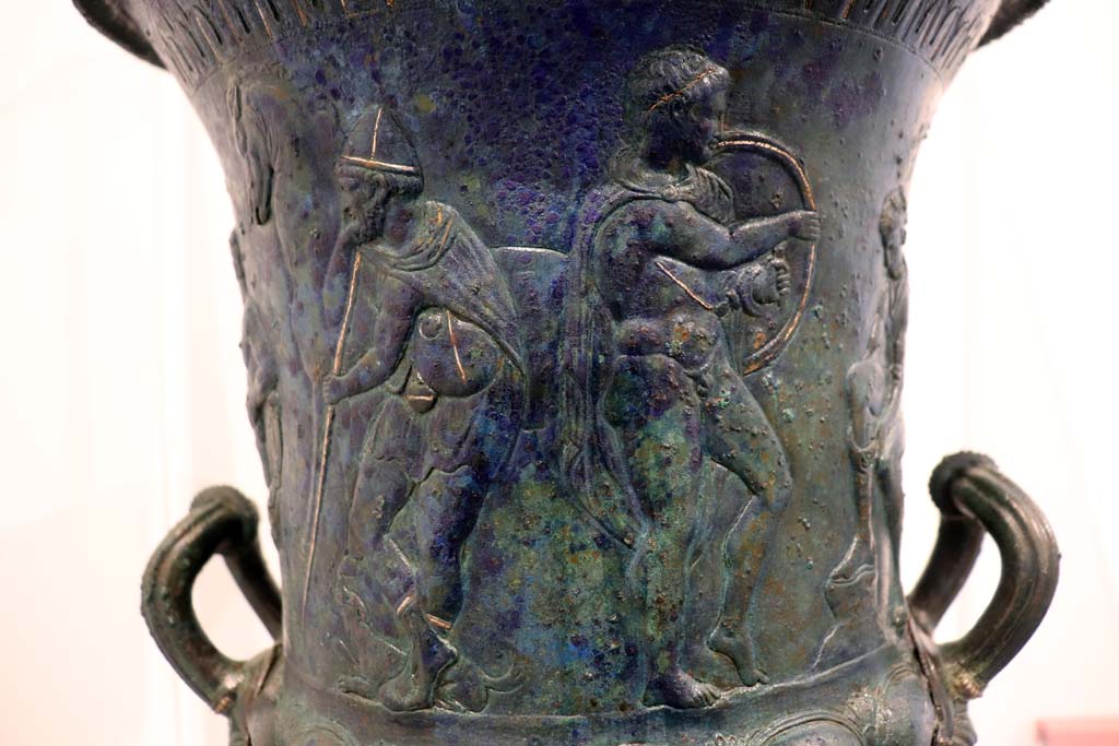 IX.13.1-3 Pompeii. February 2021.
Detail from bronze krater with scenes in relief, found in triclinium, on display in Antiquarium, VIII.1.4.
Standing in front of the seated figure tying his shoelaces are a young man holding a shield, with a bearded figure holding a staff behind.
Photo courtesy of Fabien Bièvre-Perrin (CC BY-NC-SA).