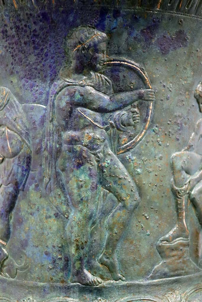 IX.13.1-3 Pompeii. February 2021.
Bronze krater with scenes in relief, found in triclinium, on display in Antiquarium, VIII.1.4.
Detail of the young man holding a shield. Photo courtesy of Fabien Bièvre-Perrin (CC BY-NC-SA).