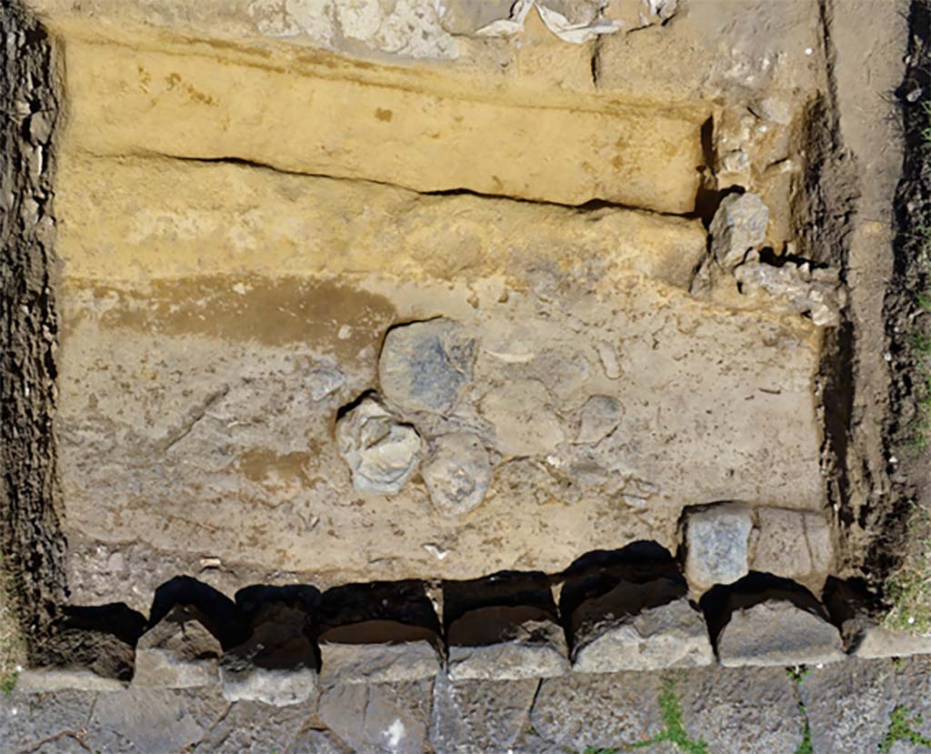 The oldest basalt road uncovered between the portico and the current paved road
Photo B. Lemaire, CJB CNRS/EFR - CC BY-NC-SA 4.0
We observed in two holes the remains of an older basalt road, lying at a lower level than the current one via dei Sepolcri, and having a slightly different orientation. 
The negative trace left by the blocks of the sidewalk shows a fairly clear difference between the ancient route and the current road.

La route en basalte la plus ancienne mise au jour entre le portique et l’actuelle voie dallée. 
Cliché B. Lemaire, CJB CNRS/EFR - CC BY-NC-SA 4.0
Nous avons pu observer dans deux sondages les restes d’une voie en basalte, plus ancienne, se trouvant à un niveau plus bas que l’actuelle via dei Sepolcri, et ayant une orientation légèrement différente. 
La trace en négatif laissée par des blocs du trottoir permet d’observer une différence assez claire entre le tracé antique et l’actuelle voie.

Pompéi : Le secteur de la Porte d’Herculanum à Pompéi : Entre espace funéraire et commercial : organisation, gestion et transformations d’une zone suburbaine 
https://centrejeanberard.cnrs.fr/spip.php?article107&lang=fr

