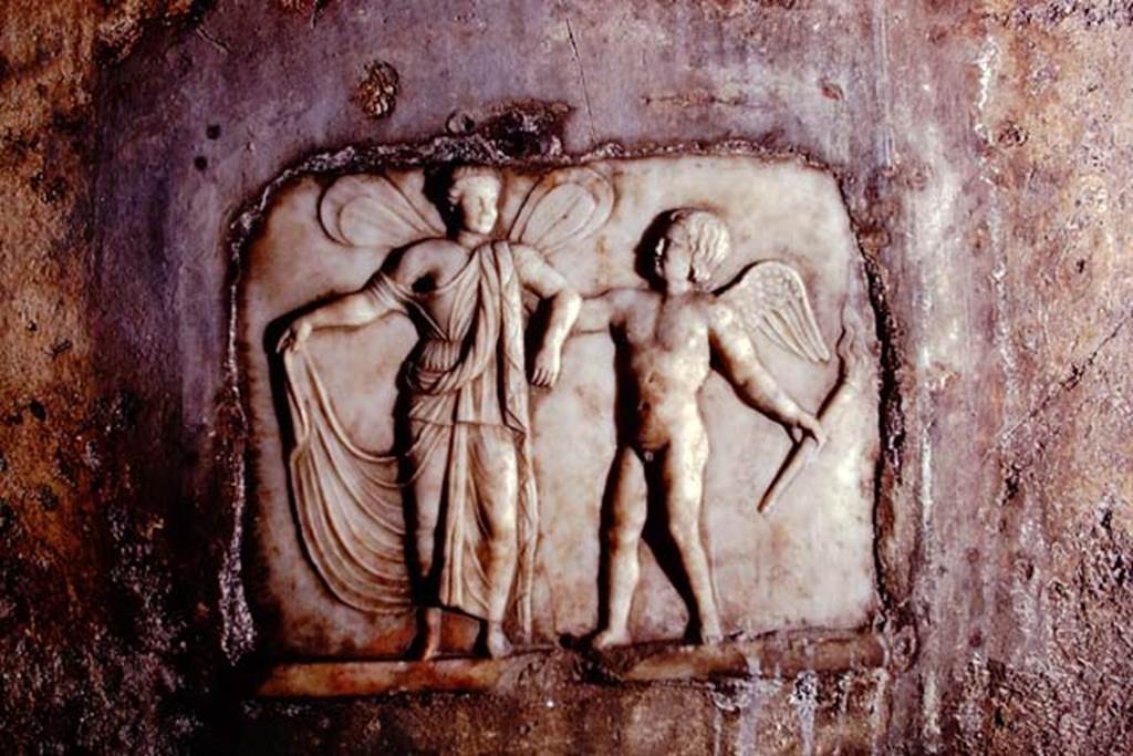Santa Maria Capua Vetere, Mithraeum. 1974. South wall, white marble relief of Eros and Psyche. 
Photo by Stanley A. Jashemski.   
Source: The Wilhelmina and Stanley A. Jashemski archive in the University of Maryland Library, Special Collections (See collection page) and made available under the Creative Commons Attribution-Non-Commercial License v.4. See Licence and use details.
J74f0582
The white marble relief (H. 0.32 Br. 0.39) was found in a recess in the southern wall above the water-basin of the podium. 
The nude winged child Amor seizes the arm of the bigger Psyche, also winged, to lead her away in the light of his torch. The girl lifts a part of her diaphanous dress. The whole is set in a red frame.
See Vermaseren M. J., 1971. Corpus Inscriptionum et Monumentorum Religionis Mithriacae, p. 108, no. 186, fig. 56.
See Minto A., in Notizie degli Scavi di Antichità, 1924, p. 367; fig. 9.