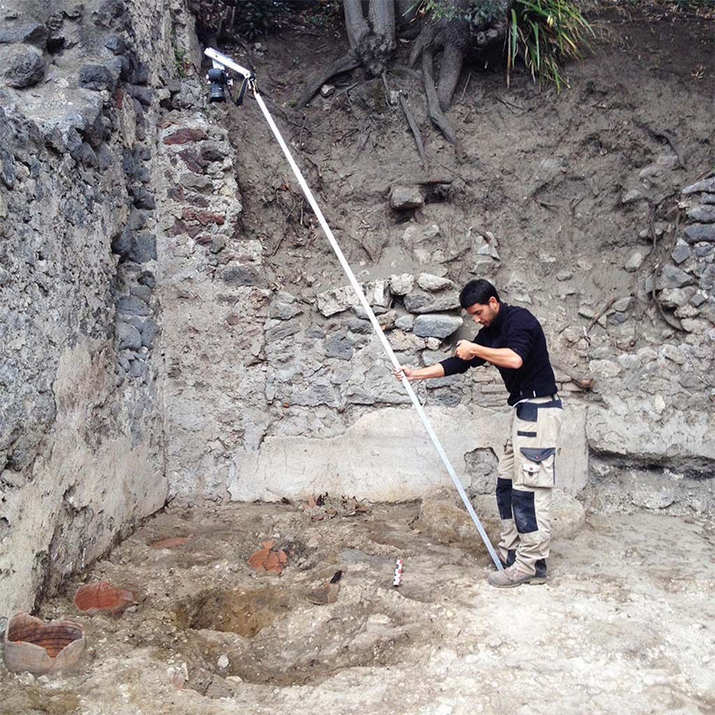HGE28 Pompeii. 2014. Bastien Lemaire photographs the locations of potter’s wheels using a pole. Photo © Guilhem Chapelin.
According to Laetitia Cavassa, after removing the modern rubble, four circular cavities were unearthed, three of whose walls consisted of fragments of cut amphoras; these were the remains of the potter’s wheels. This was a major discovery, as it illustrated through archaeological remains what we already knew, but only with regard to frescoes. Among the rare representations from the Roman period illustrating potter’s wheels at work, two are mural paintings discovered in Pompeii. Now we have these Pompeian wheels. While these traces are fleeting, they indicate the location of the master turners as well as the how these wheels worked. We noted the presence of a central axis made of wood on which the turning wheel was placed. The entire chain of operation has now been illustrated, from the shaping of vases, unfired goblets, and the firing of vases in kilns.
See Laetitia Cavassa, 2019. Tracing Back the Potters of Pompeii. Article in English
HGE28 Pompeii. 2014. Bastien Lemaire photographie les emplacements des tours de potiers à l’aide d’une perche. Photo © Guilhem Chapelin.
Rapidement, après avoir enlevé les déblais modernes, nous mettons au jour quatre cavités circulaires dont la paroi est constituée, pour trois d’entre elles, par des fragments d’amphores taillées : il s’agit des vestiges des tours de potiers dont nous cherchions les emplacements. Nous les avons enfin trouvés.
La découverte est de taille : elle vient illustrer par des traces archéologiques ce que nous ne connaissions jusqu’alors que par des fresques. Parmi les rares représentations d’époque romaine illustrant des potiers en train de travailler, deux sont des peintures murales découvertes à Pompéi. Maintenant, nous avons les tours pompéiens. Ces traces, bien que fugaces, indiquent la position des maîtres-tourneurs ainsi que le fonctionnement de ces tours de potiers. On constate la présence d’un axe central en bois sur lequel était placée la roue du tour. Toute la chaîne opératoire est désormais illustrée, de la mise en forme des vases, en passant par les gobelets crus, jusqu’à la cuisson des vases dans les fours.
Voir Laetitia Cavassa, 2019. Sur la trace des potiers de Pompéi. Article en français