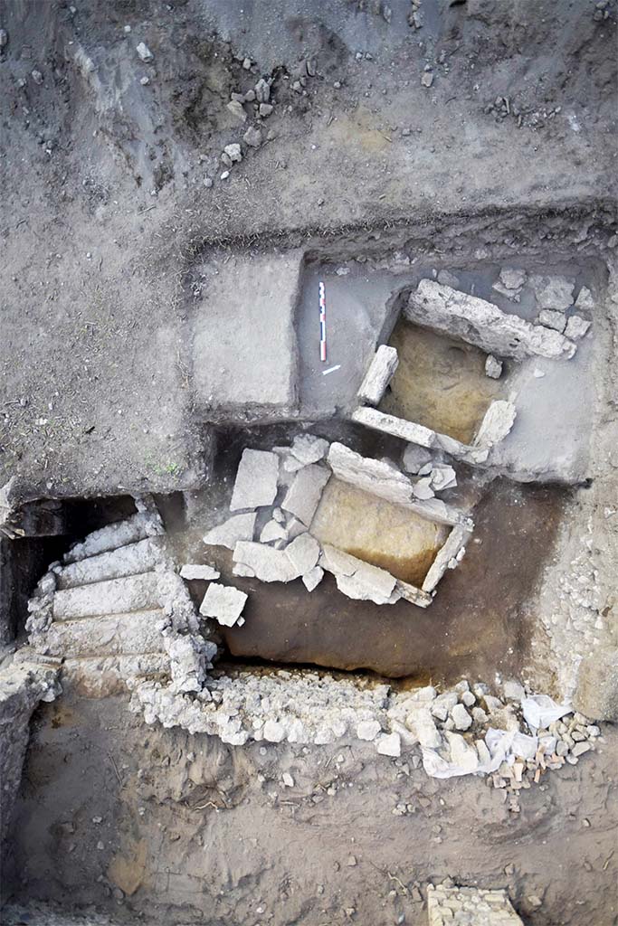 HGE31B with one side missing in 2015. HGE31A is below and the still to be uncovered HGE32A above.
HGE30 is at the bottom with the stairs of the shop at the bottom left.
Photo © B. Lemaire, EFR/CNRS.
Separated by 0.60 m from the previous one and nearly 0.50 m from the third, this grave is completely filled by reworked fill (Figs. 15 and 23). Only three Sarno limestone slabs are preserved and although the north-western one is missing, the space thus delimited is 1.44 m from north-east to south-west and nearly 0.75 m restored from north-west to south-east. The slabs measure between 0.92 and 0.99 m in height (up to 32.30 m s.l.m.) and only the one in the north-east measures only 0.62 m, but is set on wedge stones of about 10 cm. (30102).
This burial structure was excavated in the modern period, as evidenced by the levels of fill found inside. It is only a single fill. Finally, the space observed was over-excavated by our predecessors, deeper than the installation levels, making it possible to observe the foundation of the slabs and even the wedging elements of one of them. No original floor and fill levels could be observed, nor any elements relating to the remains of the deceased or to offerings.
See CEFR 1581 or para 46 and 47 below