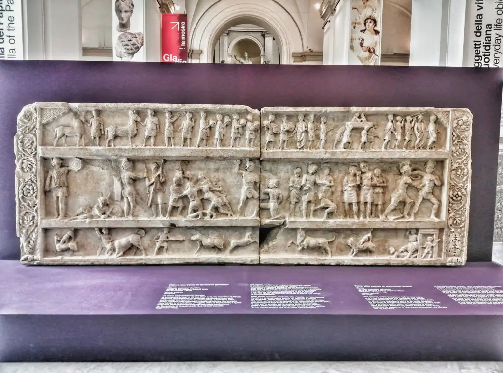 SG6 Pompeii. 2021. Gladiatorial relief with hunting scenes from a tomb at Porta Stabia. The background gives an indication of its large size.
According to Emmerson, this relief measures more than 4 metres long and 1.5 metres high.
The relief shows in three registers from top to bottom a procession, gladiator combat, and a venatio, or staged animal hunt. If this relief is funerary in nature, it is the largest known from Pompeii. 
See Emmerson A. L. C., 2010. Reconstructing the Funerary Landscape at Pompeii's Porta Stabia, Rivista di Studi Pompeiani 21, p. 78, fig. 1.
See Emmerson A., 2010. Reconstructing the Funerary Landscape at Pompeii's Porta Stabia: Rivista di Studi Pompeiani 21, pp. 78, fig. 1.
According to Osanna, the relief is in fact compatible with this monument SG6, as its length is about 4 metres long and it was found out of place, precisely in the area of Porta Stabia.
Now in Naples archaeological Museum. Inventory number 6704.

SG6 Pompeii. 2021. Rilievo gladiatore con scene di caccia da una tomba a Porta Stabia. Lo sfondo dà un'indicazione delle sue grandi dimensioni.
Secondo Emmerson, questo rilievo misura oltre 4 metri di lunghezza e 1,5 metri di altezza. Il rilievo mostra in tre registri dall'alto verso il basso una processione, un combattimento di gladiatori e una venatio, o una caccia agli animali messa in scena. Se questo rilievo è di natura funeraria, è il più grande conosciuto da Pompei.
Vedi Emmerson A., 2010. Reconstructing the Funerary Landscape at Pompeii's Porta Stabia: Rivista di Studi Pompeiani 21, pp. 78, fig. 1.
Secondo Osanna, il rilievo è infatti compatibile con il monumento SG6, in quanto la sua lunghezza è lunga circa 4 metri ed è stato trovato fuori luogo, proprio nella zona di Porta Stabia.
Ora al Museo archeologico di Napoli. Numero di inventario 6704.

