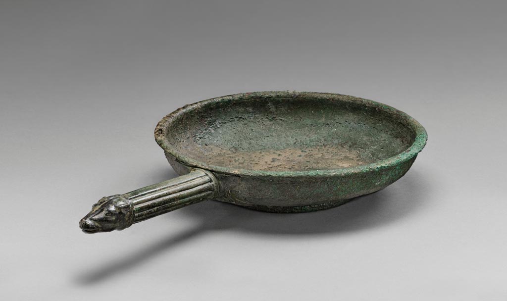 Discovered in a villa in Boscoreale. Bronze patera with dog’s head handle.
Digital image courtesy of the Getty's Open Content Program. Now in the Getty Museum, inventory number 72.AC.134.
The interior and exterior of this bronze patera (shallow bowl) are undecorated. A series of concentric bands decorate the underside of the foot. The tip of the fluted handle is fashioned in the shape of a dog’s head. At the join to the bowl is a relief attachment comprised of floral ornaments.
Vessels of this type were used in the home for religious rituals and to hold water for handwashing during banquets. Slaves would pour water from an oinochoe (pitcher) over the hands of participants, catching the cascade in a patera held underneath. This patera was discovered in a villa at Boscoreale or in the vicinity, although the exact findspot is unknown. 
