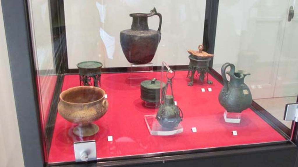 Gragnano, Villa rustica in Località Carmiano, Villa A.
Stabiae Antiquarium, inventory numbers
63464 Cup left found in room 7.
63528 bronze perfume holder (centre) found in room 1.
63437 pyx (behind) found in room 9.
63377 jug (rear) found in room 4.
63278 (left rear) 63279 (right rear) lamp stands found in room 2.
63543 lamp found in room 6.
63539 jug (front right) found in room 6.
Photo courtesy of Donna Dollings.