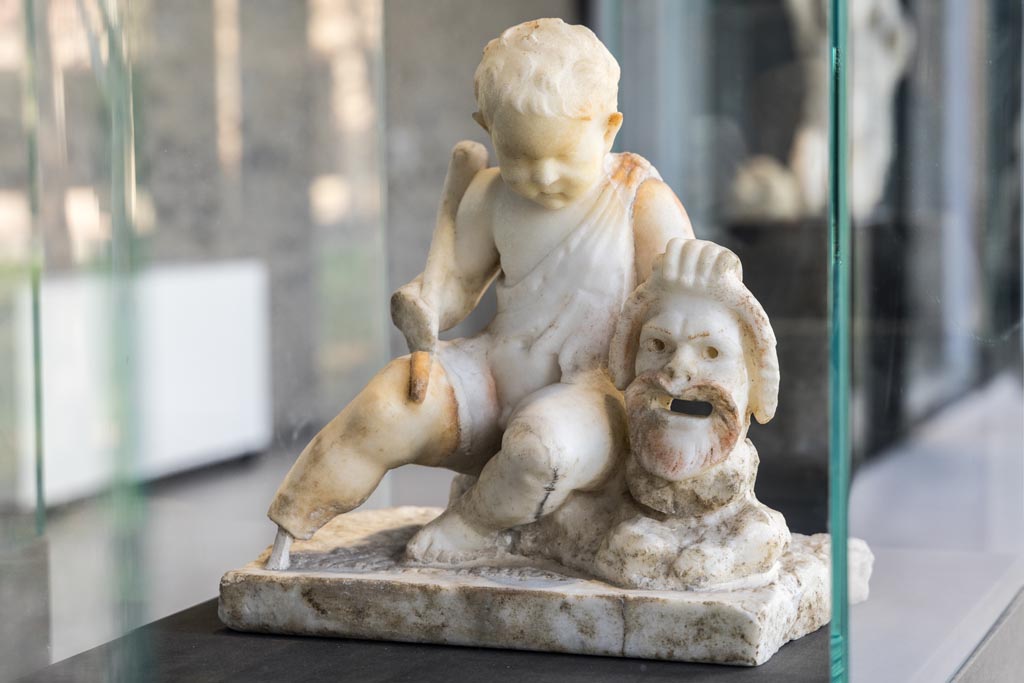 II.2.2 Pompeii. January 2023. Room “l”, (L), garden. White marble statuette of a cupid with a mask.
Found above the marble steps in the nymphaeum at the top of the lower euripus in the garden in 1920.
On display in exhibition in Palaestra. Photo courtesy of Johannes Eber.